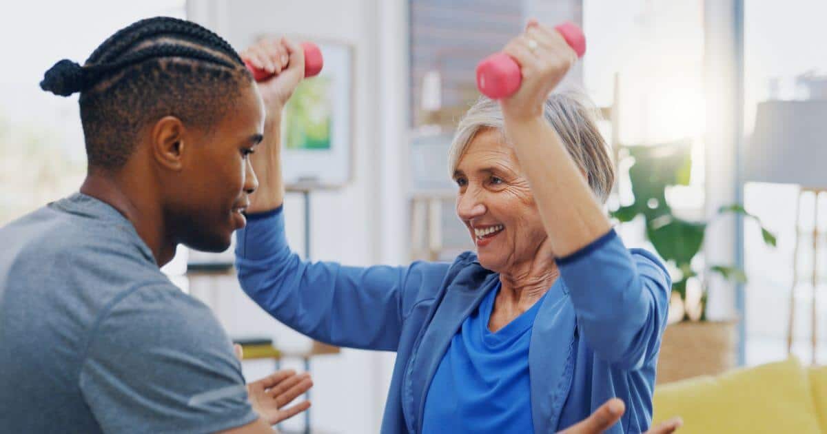 Elder woman in a fitness class for seniors lifting weights with a caregivers assistance.