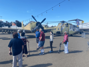 A group of older adults stands outside with a World War II-era plane and helicopter in the background