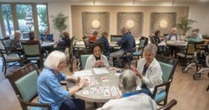 A group of four older women sit around a table playing cards, while groups of people at three tables behind them play their own card games