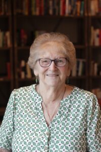 An older smiling woman in glasses and green patterned blouse in front of a bookcase