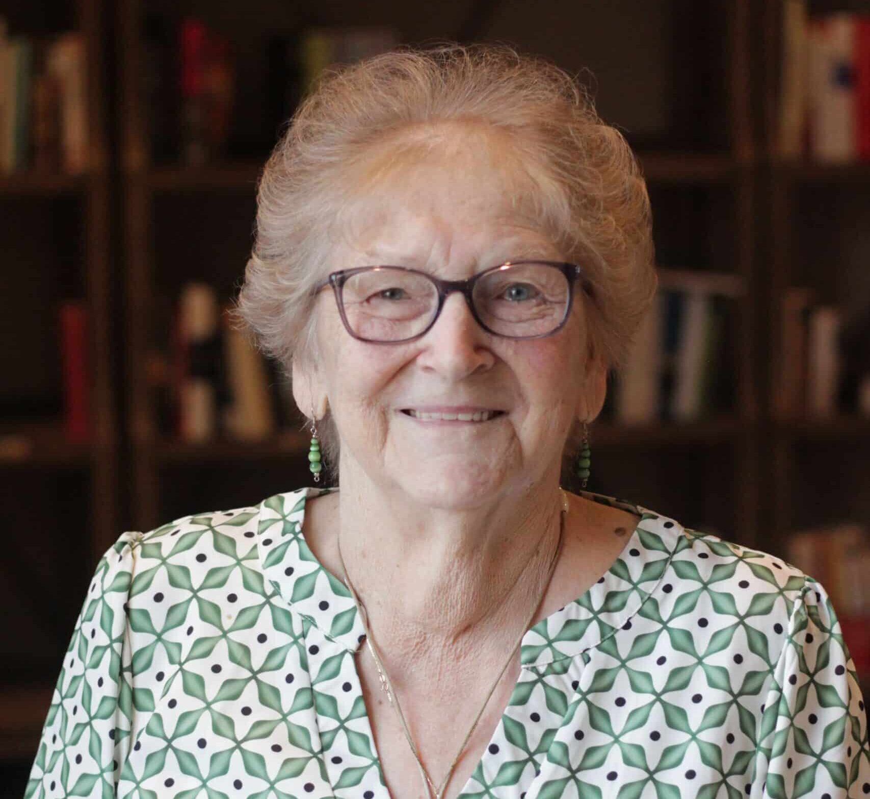 An older smiling woman in glasses and green patterned blouse in front of a bookcase