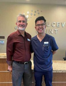 Two men standing together smiling; on the right is young man with glasses who works as a med tech wearing navy scrubs; on the left is a man in shirt and dress pants.